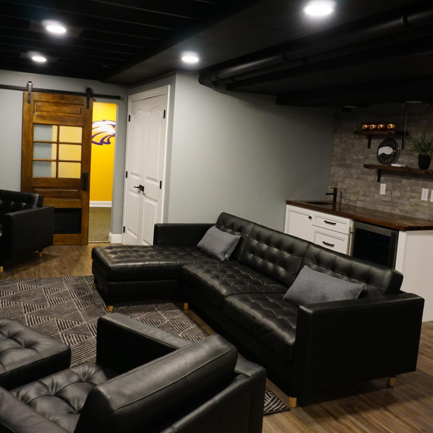 Basement living room with large black sectional, wood barn door, and small kitchenette.